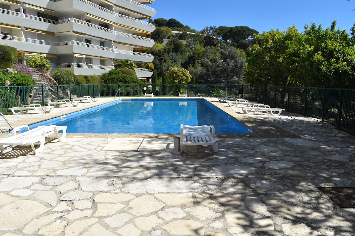 A clear blue pool is surrounded by a spacious stone area, furnished with sun loungers. Lush greenery and landscaped gardens are visible in the background, providing a serene atmosphere. The building's unique architecture complements the outdoor space.