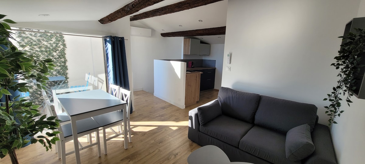 An open living area showcases a modern kitchen adjacent to a cozy seating space. White walls and wooden beams create an airy atmosphere. A dining table with chairs is positioned beside a window, enhancing natural light. Simple greenery decorates the room.