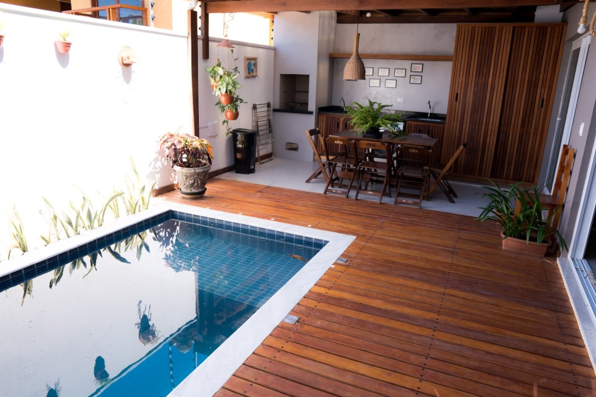 A wooden deck surrounds a calm pool, reflecting the surrounding greenery. A dining area with wooden chairs is set near a kitchen space, while potted plants add a touch of nature. The area is designed for relaxation and outdoor dining.