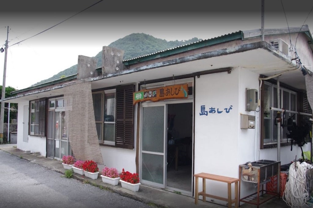 The exterior of a low-profile building is exhibited, featuring large windows and a welcoming entrance. Potted flowers are seen lining the pathway, adding a touch of color. A sign with the name "島あしび" is prominently displayed, indicating the establishment's identity.