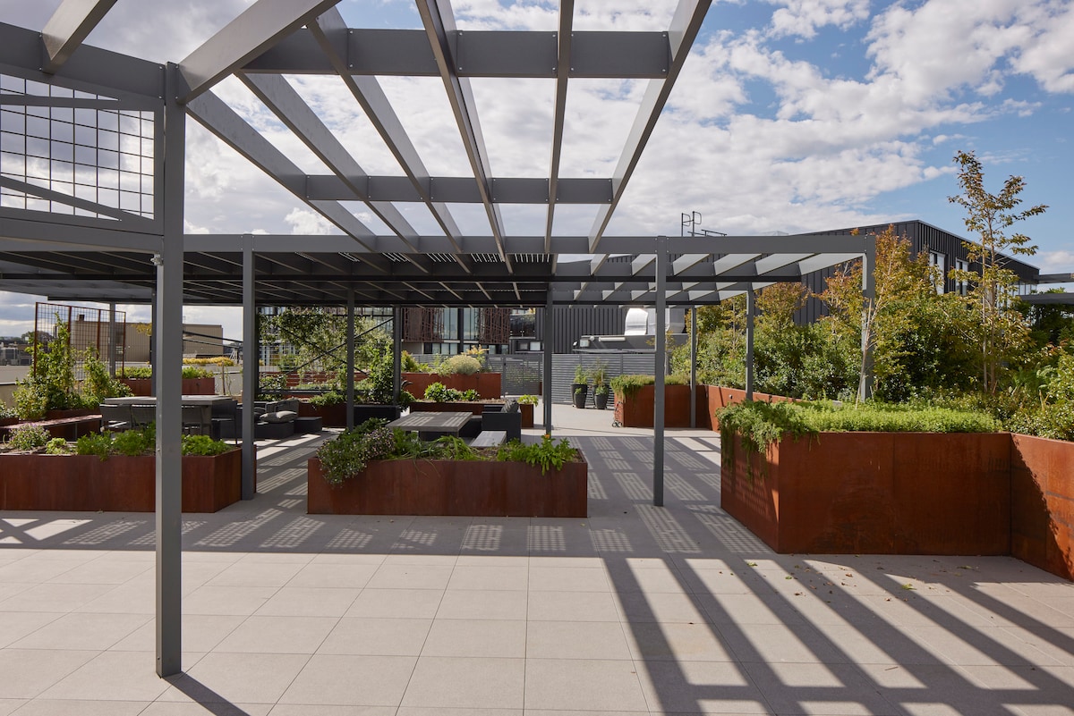 A spacious rooftop area is surrounded by planter boxes filled with greenery. An open canopy provides shade over seating arrangements, with textured gray flooring leading through the space. Clear blue skies and distant buildings create a contrasting backdrop for this outdoor relaxation area.