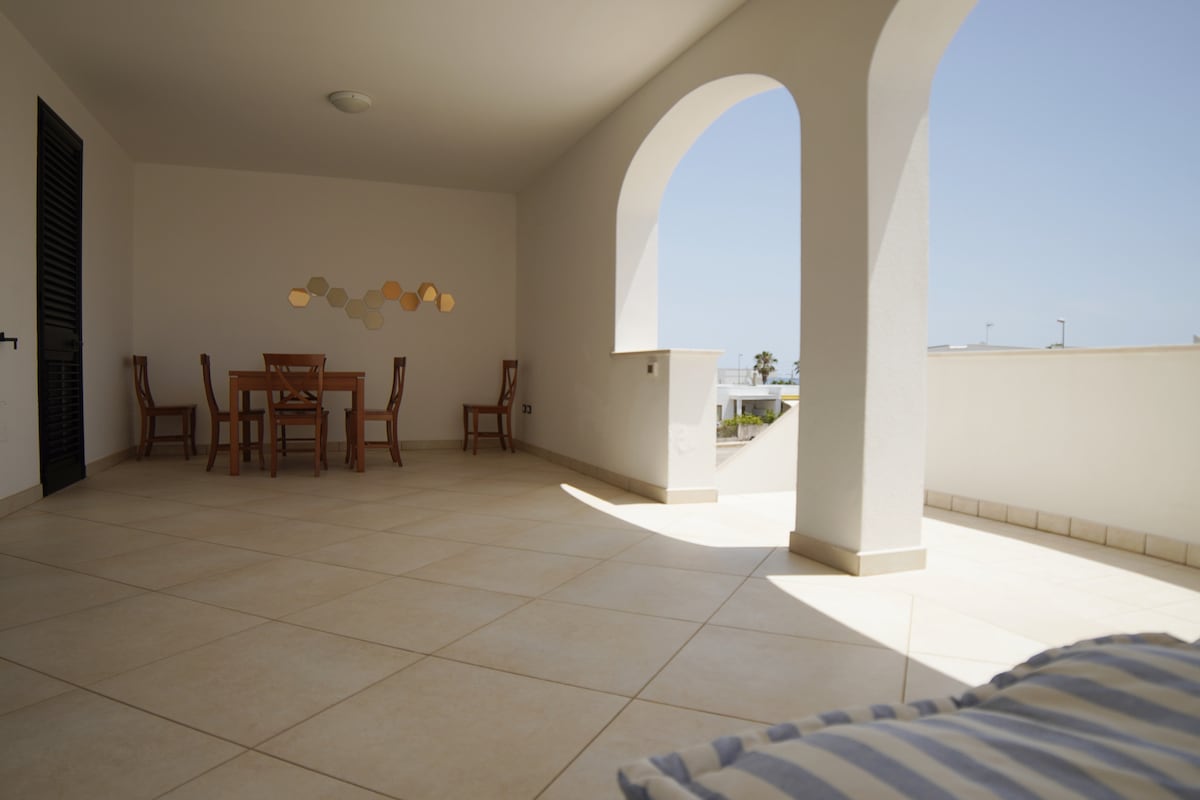 A spacious terrace is visible, featuring a dining table surrounded by six chairs. Natural light fills the area through an arched opening, and the tiled floor enhances the open feeling. Light-colored walls create a bright atmosphere, with a simple decorative element on the wall.