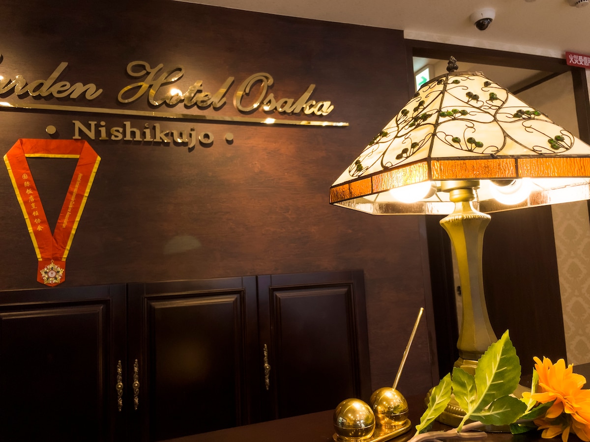 The reception area features a decorative lamp with an intricate floral design, casting a warm glow. A wooden sign displaying the hotel name is mounted on the wall. Elegant accessories, including a golden decorative piece and green leaves, add a refined touch to the welcoming space.