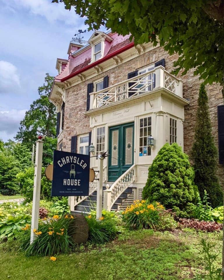 The exterior of Chrysler House features a stately stone facade adorned with intricate architectural details. A welcoming sign is positioned near the entrance, surrounded by vibrant greenery and colorful flower beds, enhancing the charming appeal of this historic residence.