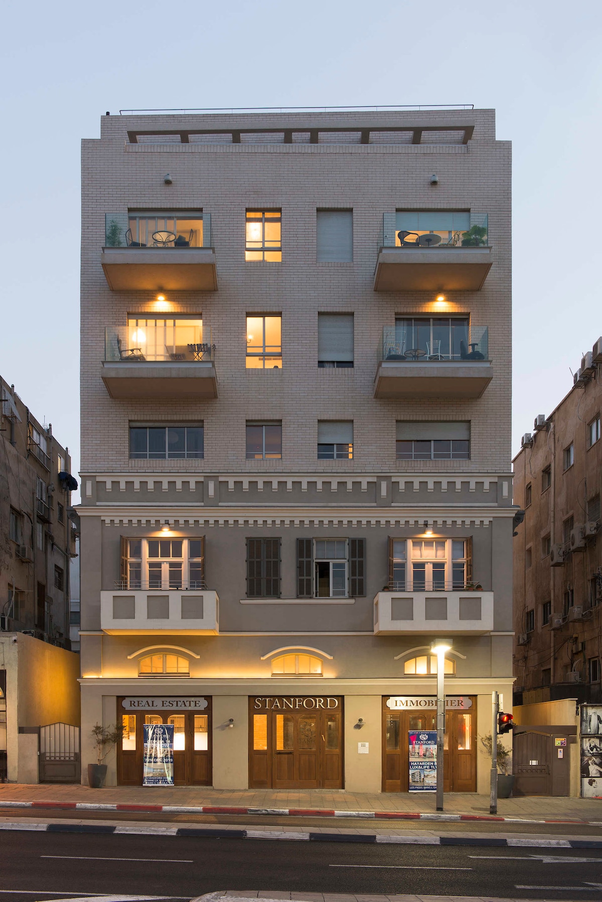 The building features a striking eclectic façade with a mix of old and new architectural elements. Illuminated balconies enhance the design, while warm lighting highlights the entrance. Street-level commercial spaces are showcased, contributing to the vibrant atmosphere of the neighborhood.