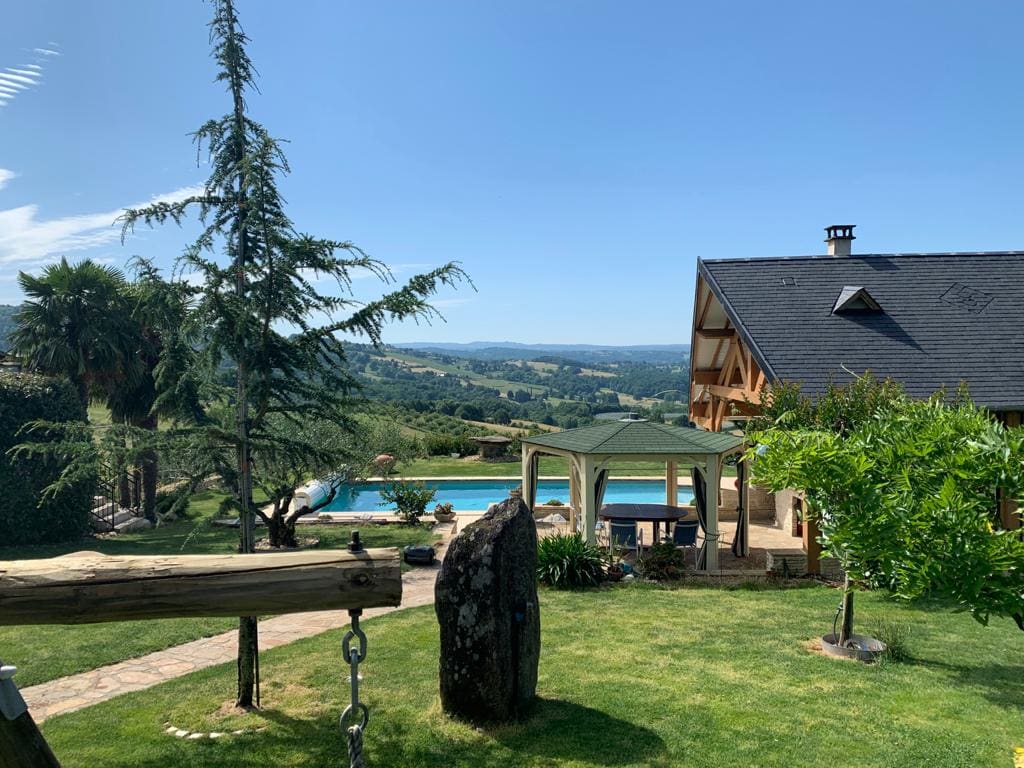 A spacious outdoor area is depicted, featuring a large swimming pool surrounded by lush greenery. In the background, a beautifully crafted wooden house is visible. A shaded seating area is set up near the pool, with distant scenic hills creating a serene backdrop.