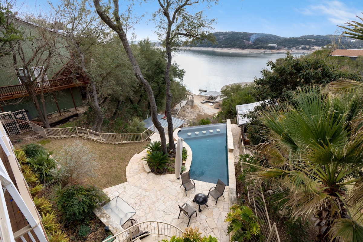 An aerial view showcases a private swimming pool with zero-edge design, positioned next to tranquil landscaped areas. The cove of Medina Lake is visible in the background, framed by trees and stone pathways leading down to the water. Outdoor seating is arranged nearby.