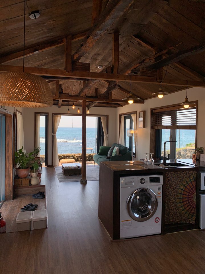 An open living area features a modern kitchen with a washing machine, beneath a rustic wooden ceiling with exposed beams. Large windows frame views of the sea, while natural light illuminates the space. A comfortable seating area is positioned to enjoy the ocean scenery.