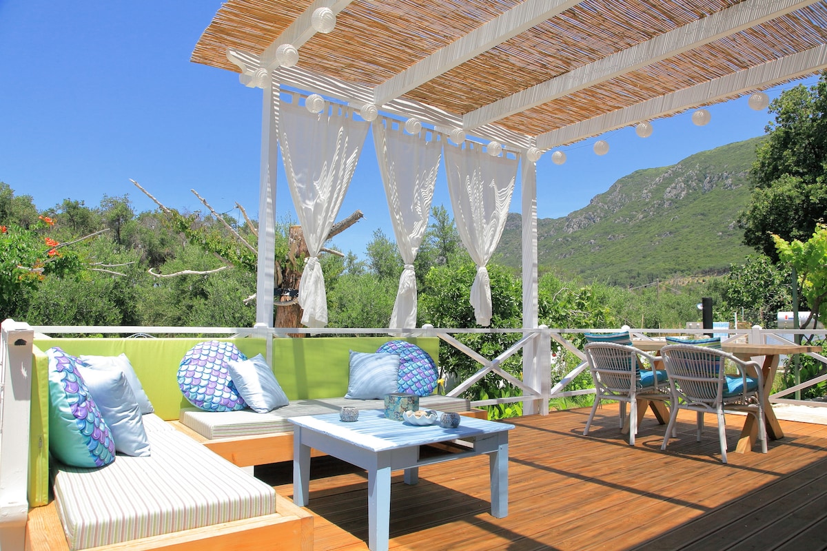 An inviting outdoor deck features comfortable seating with green cushions, white tables, and decorative pillows. Sheer curtains gently sway in the breeze, while lush greenery and hillside views provide a serene backdrop. Soft lighting accents are positioned along the pergola for evening ambiance.