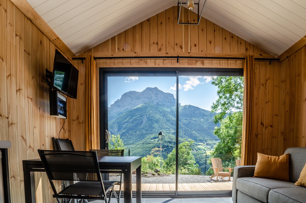 A wooden interior features large glass doors that offer a panoramic view of the surrounding mountains. A dining table with chairs is positioned near the window. A cozy seating area includes a gray sofa and a small side table, while natural light fills the space through the open doors.