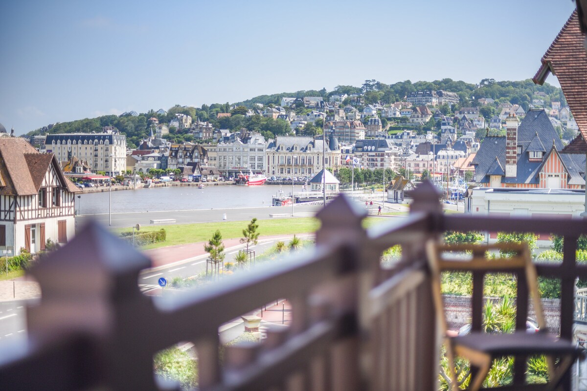 A view from the balcony captures the waterfront, featuring a marina lined with boats and charming buildings dotting the hillside. The scene includes green hills in the background, enhancing the coastal environment, with clear skies above.