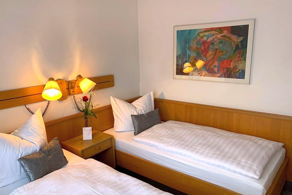 A bright guest room features two separate beds with crisp white bedding and decorative cushions. A bedside table holds a small vase with a rose. Soft lighting is provided by wall-mounted lamps. A colorful framed artwork adorns the wall above the beds.