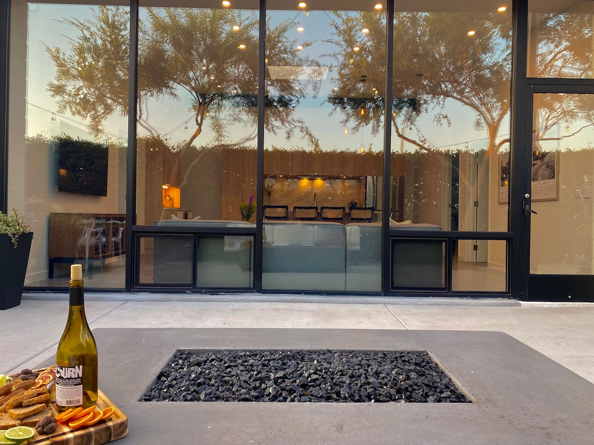 A modern patio showcases an oversized fire pit surrounded by black gravel. A bottle of wine is placed next to a platter of snacks, with large glass doors reflecting the interior space. Trees are visible in the background, contributing to a tranquil atmosphere.