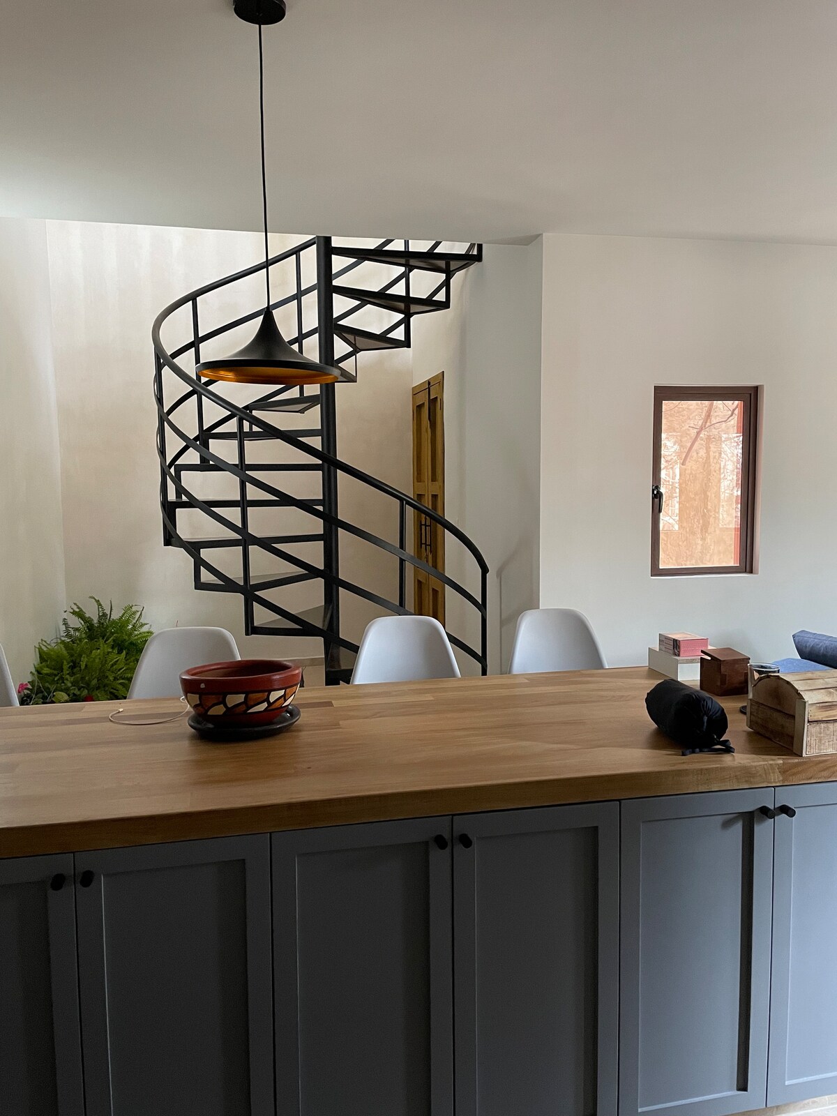 A modern kitchen area features a wooden countertop and sleek grey cabinetry. A spiral staircase made of black metal ascends to the upper floor. White dining chairs surround the table, with natural light filtering through a small window nearby.
