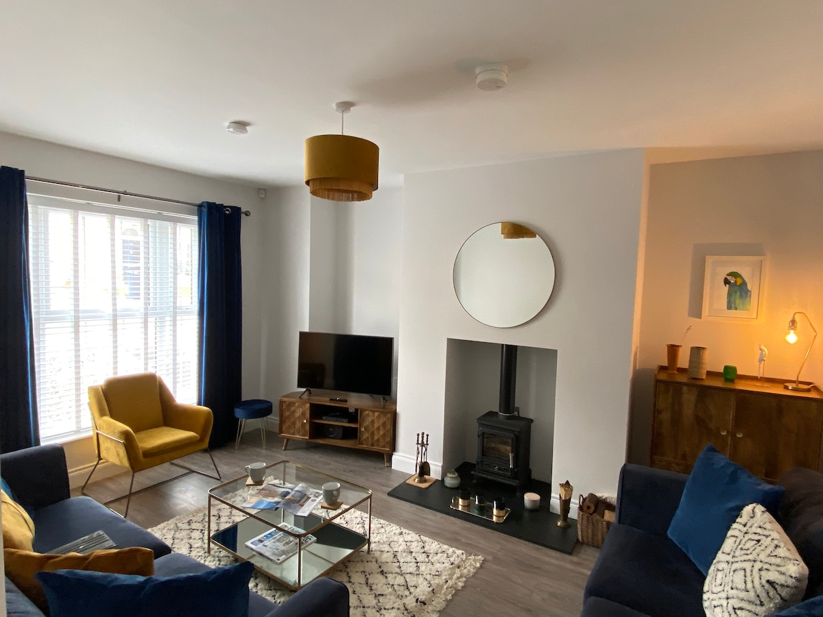 A modern lounge area is presented, featuring a yellow accent chair, a navy sofa, and a glass coffee table. Natural light filters through the window adorned with blue curtains, while a flat-screen TV is positioned near a stylish fireplace. Decorative elements enhance the welcoming space.