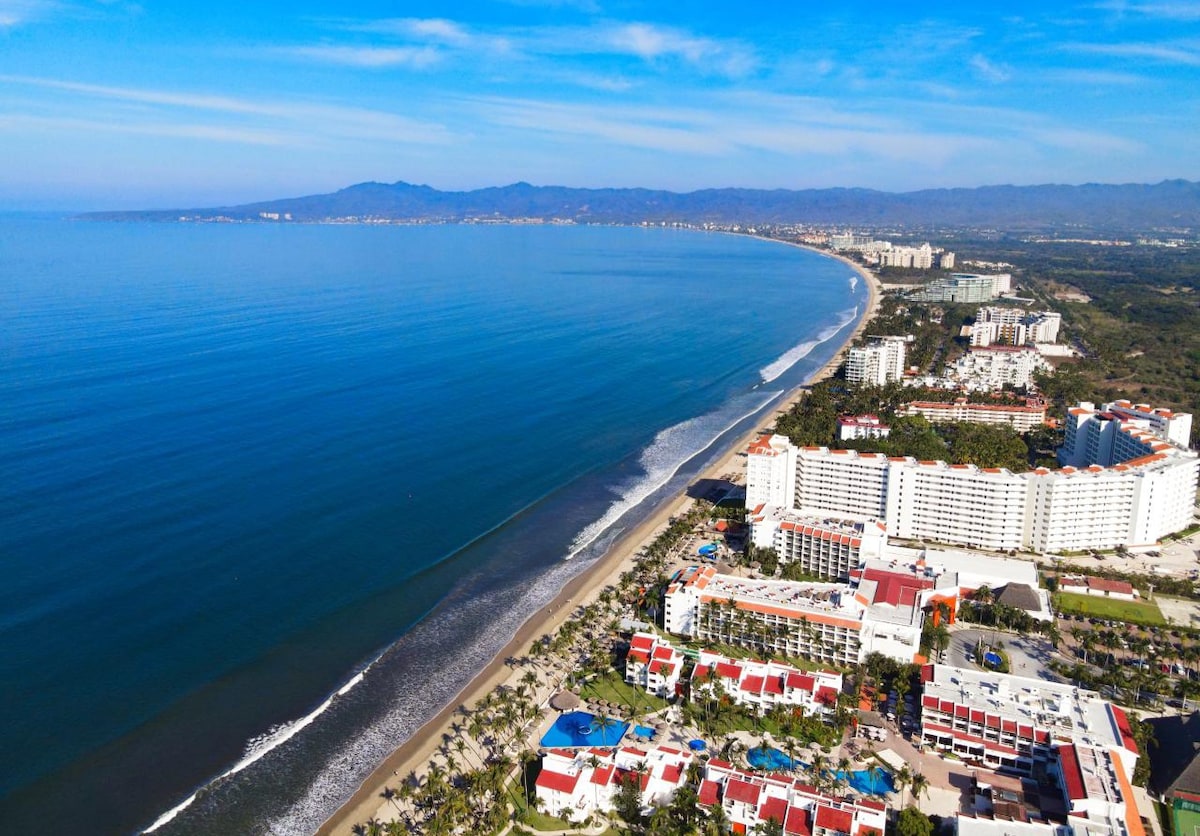 Aerial view of a coastal area showcasing a long stretch of sandy beach bordered by gentle sea waves. The mountains serve as a backdrop, and various buildings are visible along the shore, highlighting the blend of nature and urban development.