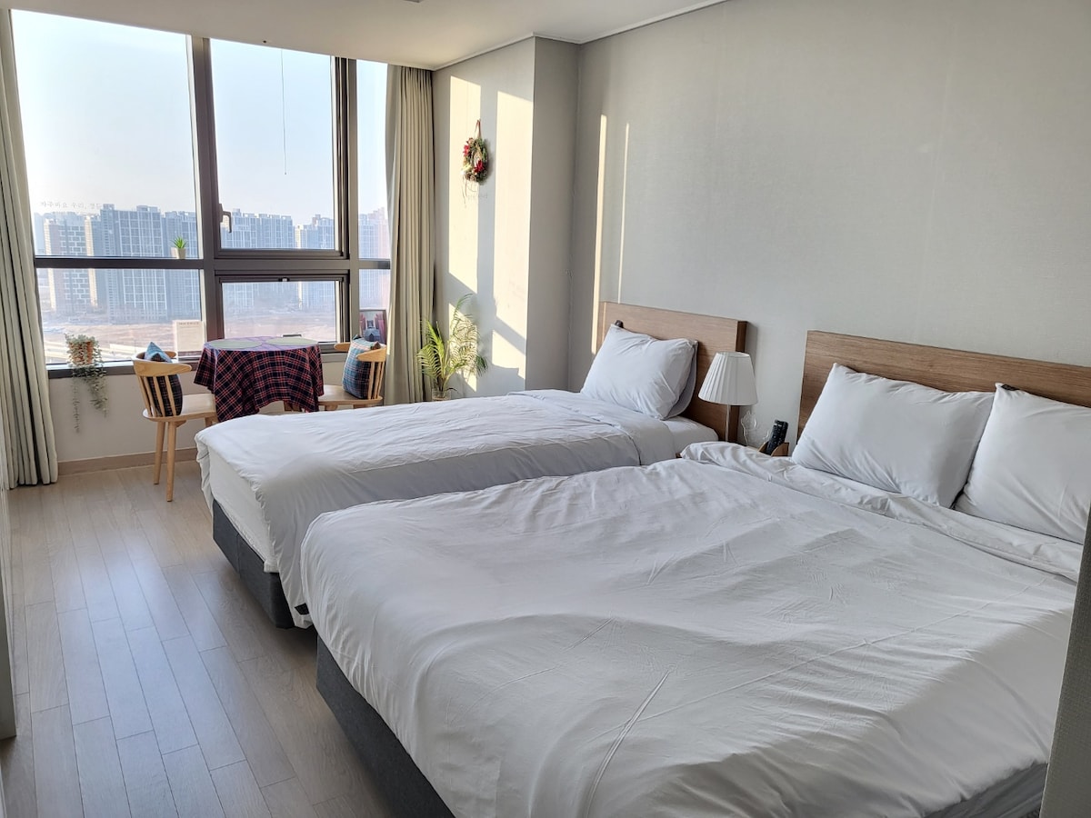 A bright and airy bedroom features two spacious beds with crisp white linens. Large windows provide abundant natural light and showcase a city view, while a small table and chair are placed near the window. Decorative plants add a touch of warmth to the space.