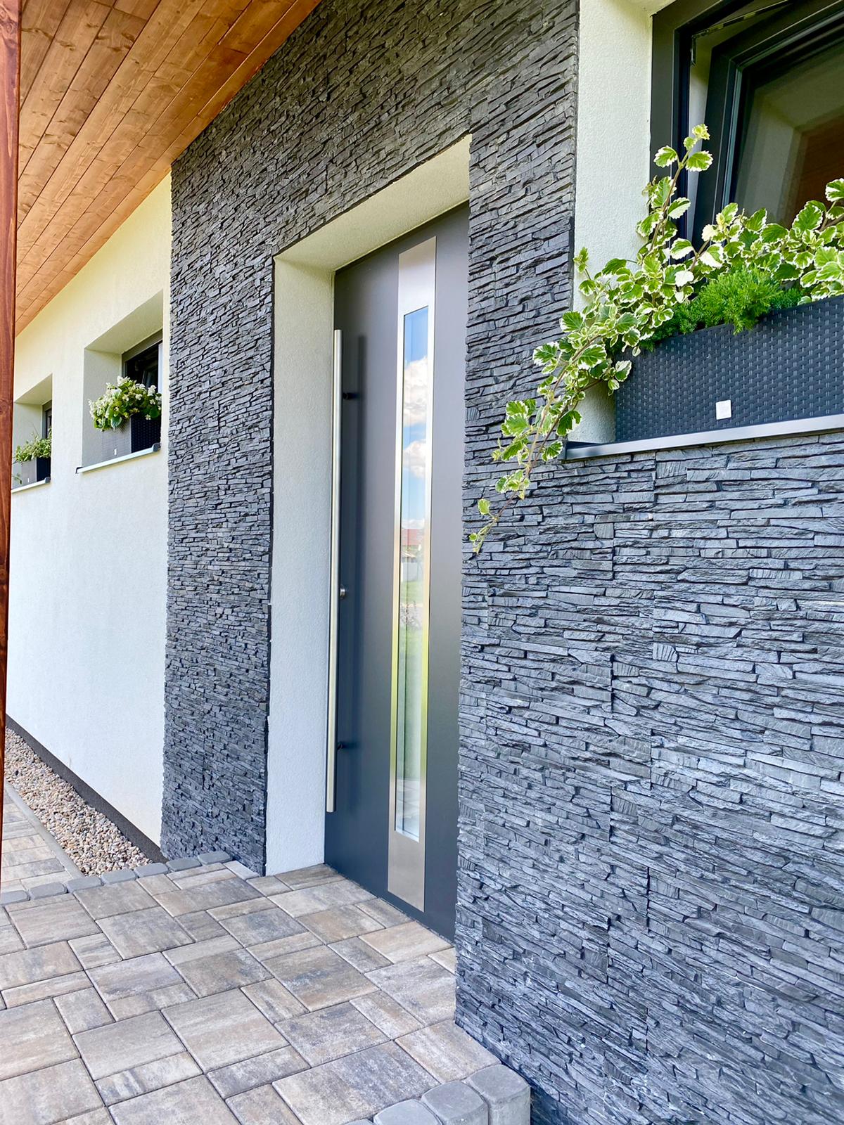 The entrance features a sleek, modern door framed by textured stone walls. Two small windows above the doorway are adorned with green plants, adding a touch of nature. A paved path leads up to the door, providing an inviting entrance.