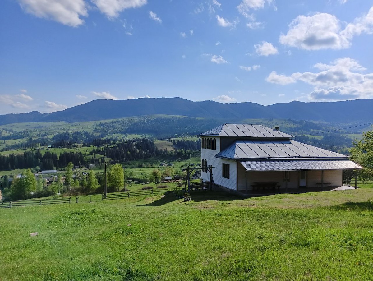 A modern two-story house is set against a backdrop of lush green hills and the distant mountain range. Sunlight brightens the exterior with a sloped roof and broad windows. Surrounding grassy areas create a spacious environment, inviting outdoor relaxation and exploration.