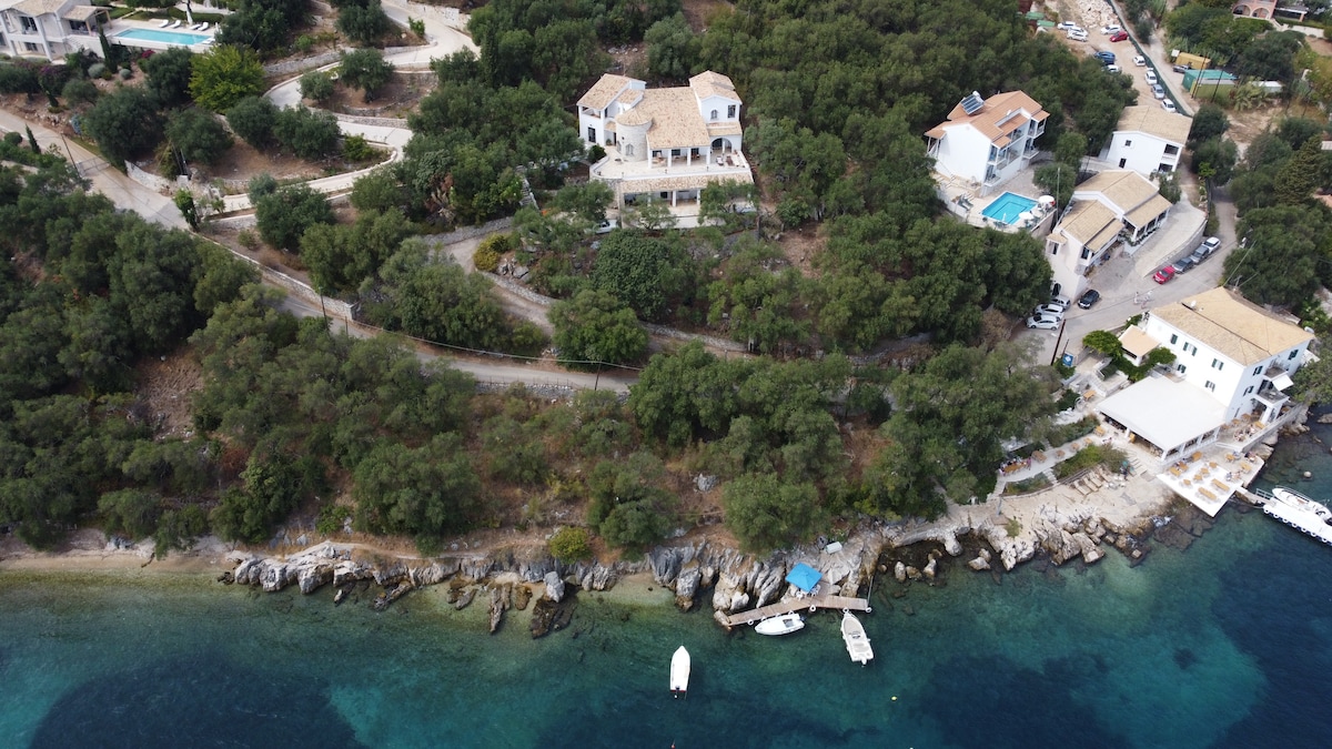 An aerial view captures a coastal landscape characterized by lush greenery and rocky shoreline. Several villas are visible along the hillside, with pathways leading towards the water. Boats are anchored in the clear blue sea, highlighting the tranquil coastal setting.