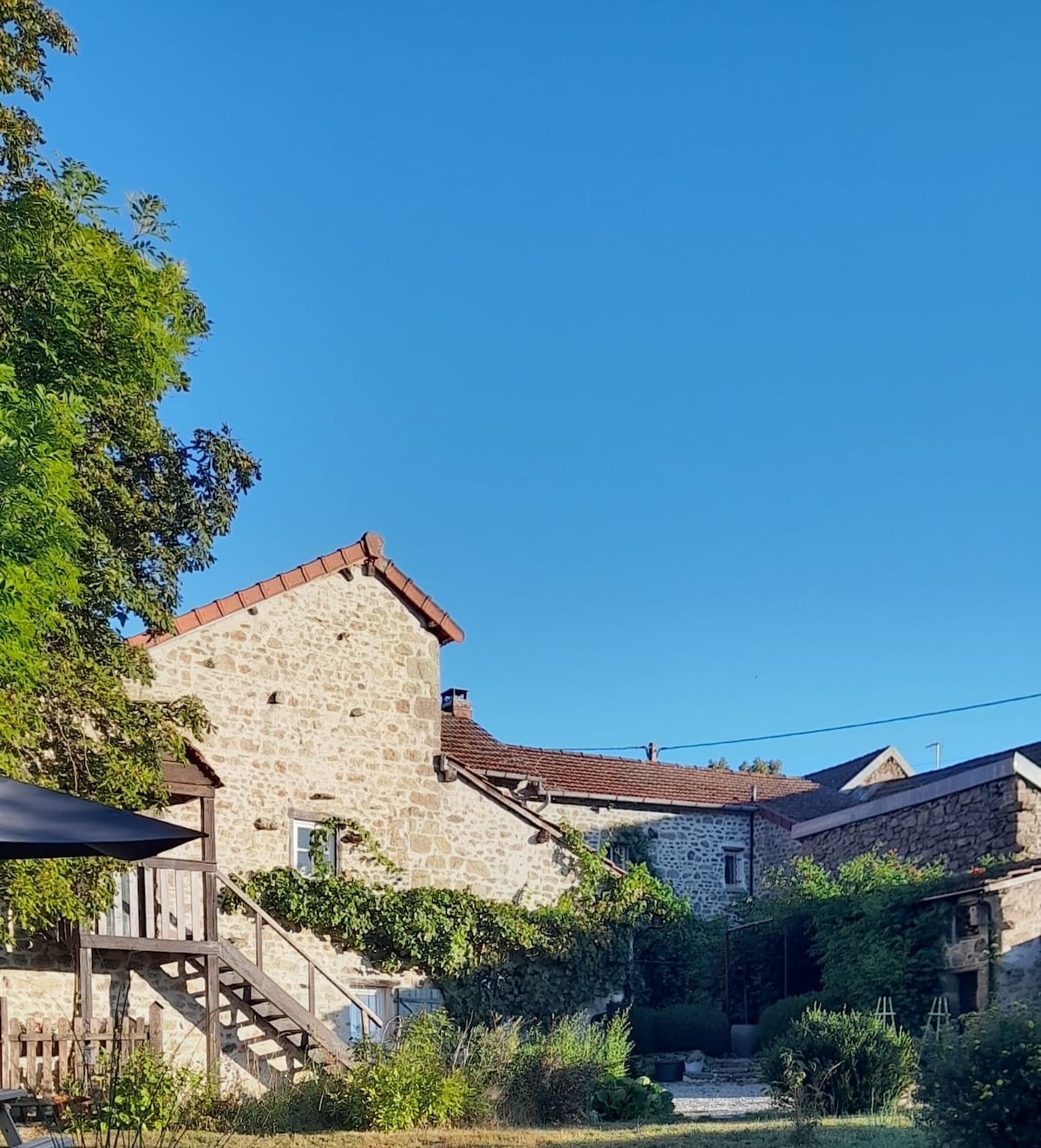 The exterior of the lodge is presented, showcasing a stone facade and a sloping roof. A staircase leads to an upper level, surrounded by green foliage. The bright blue sky creates a vibrant backdrop against the rustic architecture.