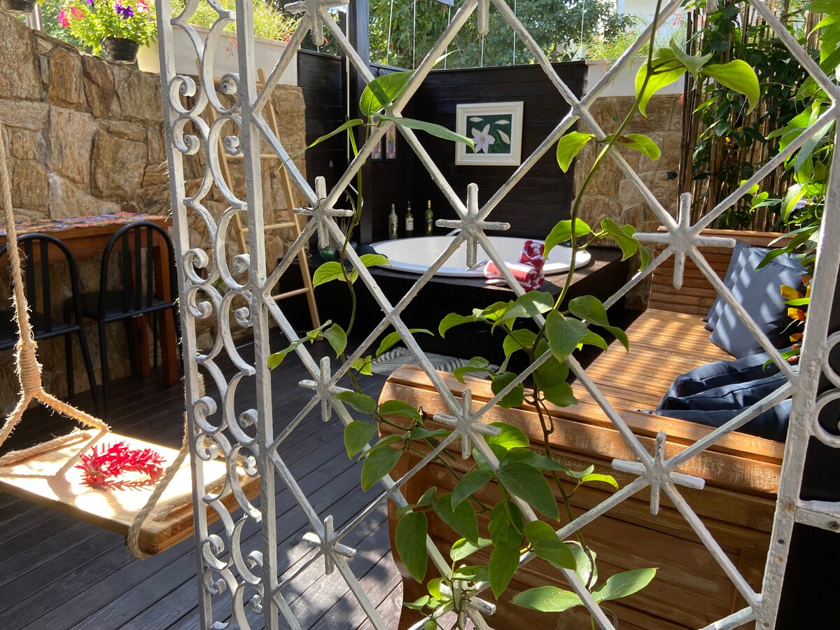 A serene outdoor area features a comfortable jacuzzi framed by wooden decking and lush greenery. A decorative iron gate adds a touch of charm, while a cozy swing provides a relaxing spot. Bottles are visible on a shelf, enhancing the tranquil environment.