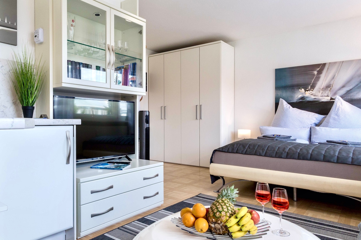 A modern studio apartment features a spacious bed with soft linens, complemented by a large wall-mounted TV. A stylish dining table displays fresh fruit and two glasses, inviting relaxation. Neutral-toned furniture enhances the contemporary decor, while natural light brightens the room.