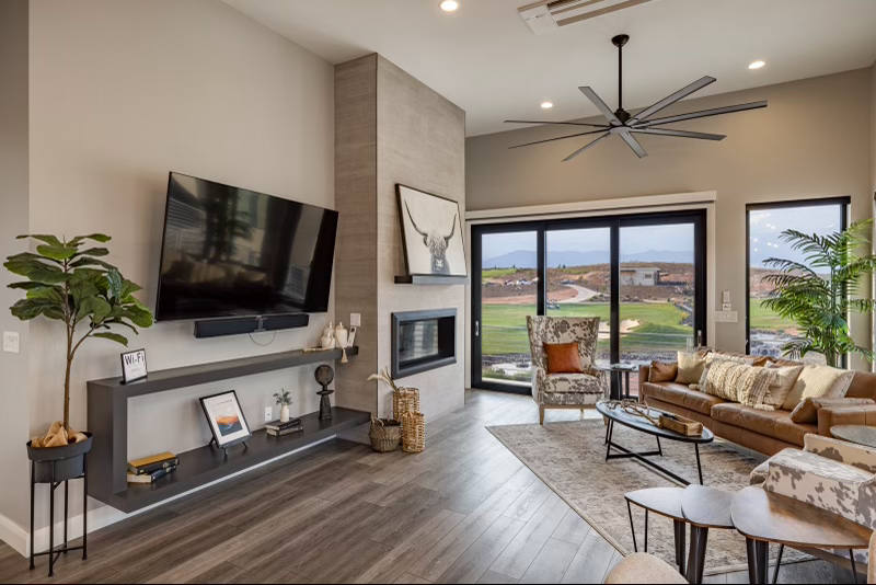 The living room features a spacious seating area with a large sectional sofa and accent chairs, offering a welcoming space for relaxation. A flat-screen TV is mounted on the wall, with a sleek entertainment console below. Large windows provide ample natural light and views of the outdoor landscape.
