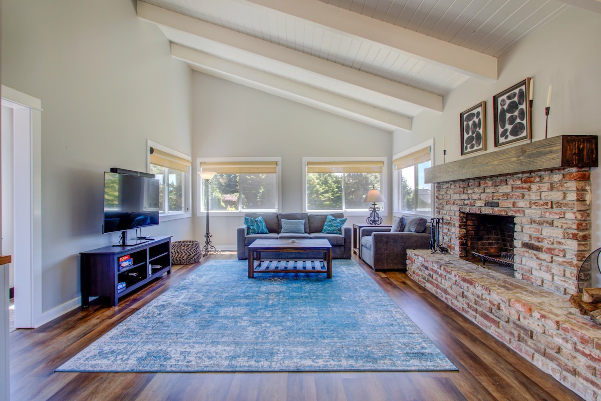 A spacious living area features a large, comfortable couch and an armchair, arranged around a wooden coffee table. Large windows allow natural light to fill the room. A stone fireplace serves as a focal point, complemented by a vibrant area rug on the wooden floor.