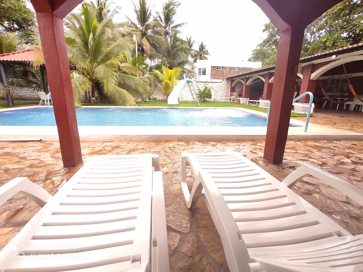 Lounge chairs are positioned by a swimming pool, which features a water slide and is surrounded by lush palm trees. The tranquil pool area is complemented by a stone patio, providing a spacious area for relaxation and enjoyment.