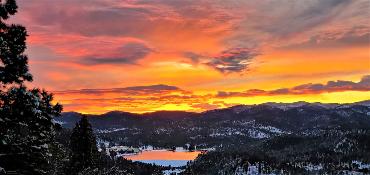A vibrant sunset casts warm hues of orange and pink across the sky, illuminating a snow-covered landscape. Rolling hills and a serene lake are visible in the foreground, framed by evergreen trees on the left, creating a picturesque natural setting.