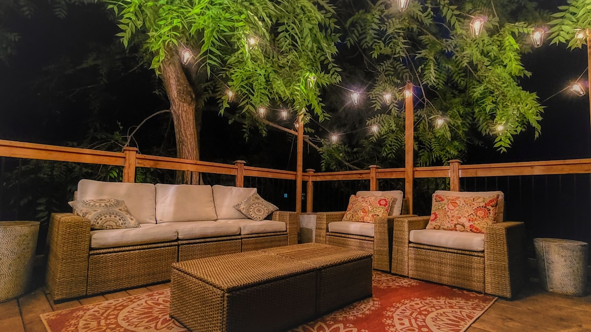 A spacious outdoor deck area is illuminated by warm string lights. Comfortable seating arrangements include a large sofa and two armchairs, all adorned with patterned cushions. A central coffee table adds functionality, while surrounding greenery enhances the inviting atmosphere of the space.