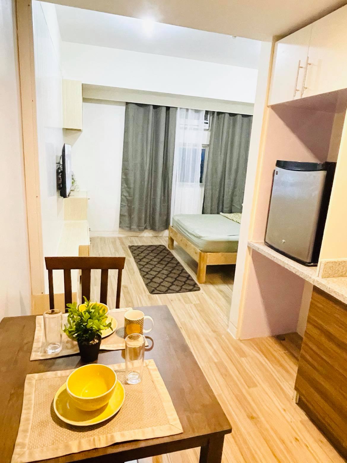 An inviting studio space is revealed, featuring a dining area with a wooden table and yellow bowls. In the background, a cozy sleeping area with a modest bed is framed by soft gray curtains, while a kitchenette and a small refrigerator are visible to the right.