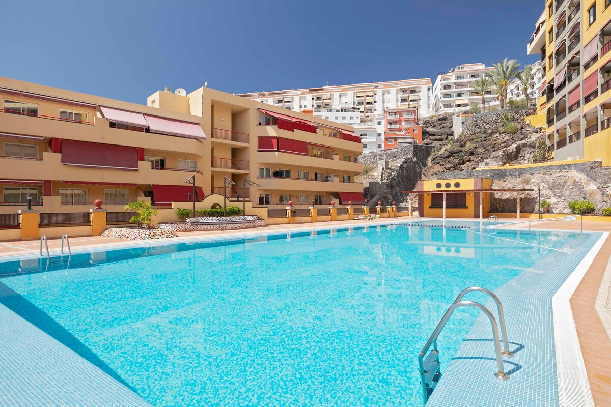 A large swimming pool is framed by a modern apartment complex. The clear blue water reflects the vibrant sunlight, with steps leading into the pool. Surrounding landscaping includes stones and plants, providing a serene atmosphere. The background features multi-story buildings and palm trees.