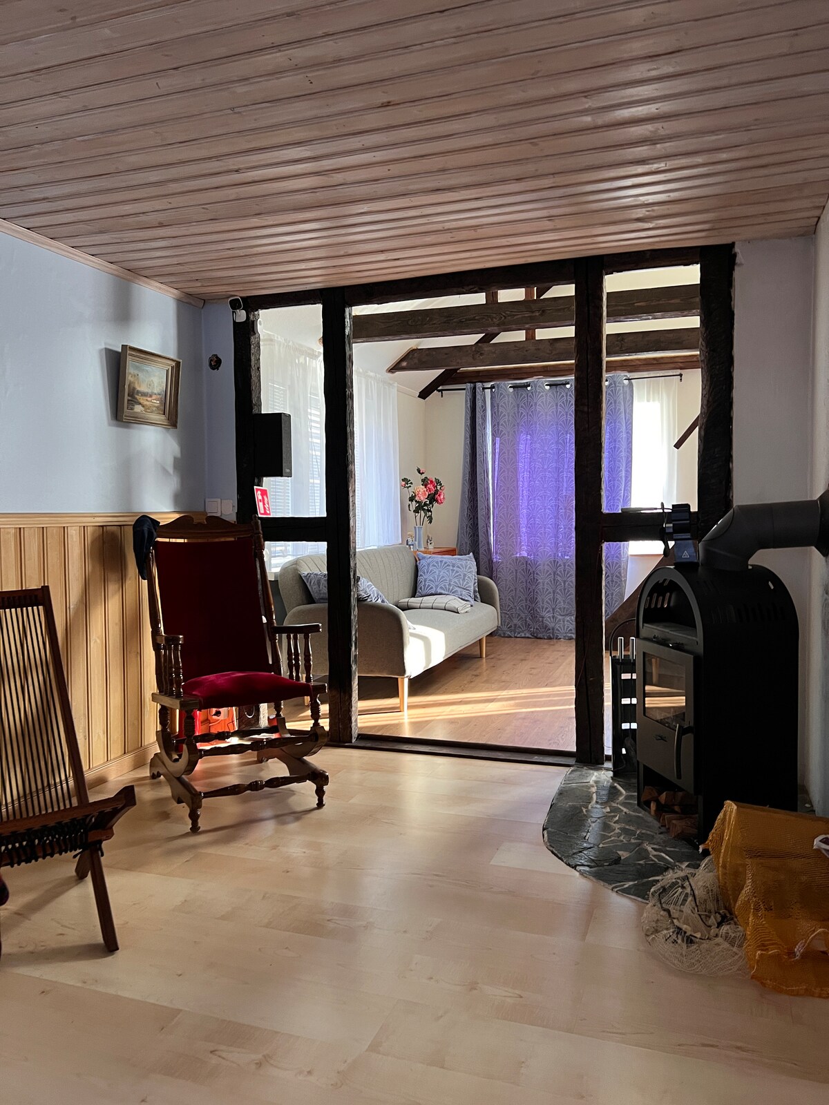 A welcoming living area is viewed through a wooden framed entry. A comfortable sofa is positioned against a wall draped with sheer curtains, allowing soft light to filter in. A decorative chair and a wood fireplace add character to the warm ambiance.