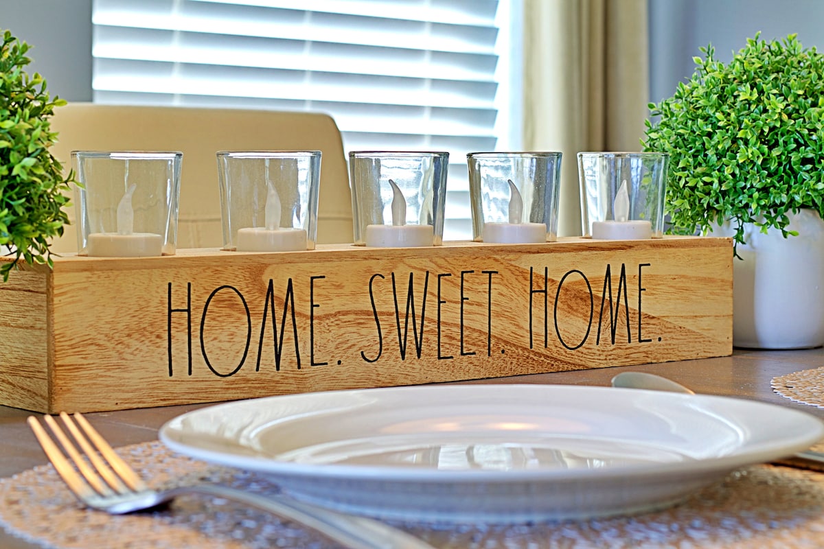 A wooden centerpiece features the phrase 'HOME. SWEET HOME.' with four glass candle holders positioned atop it. Two small potted plants are placed on either side. A decorative placemat and a white plate set on a dining table complement the setup.