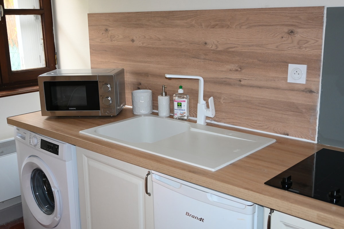 A modern kitchen space is featuring a wooden backsplash and a smooth countertop. Essential appliances are included, such as a microwave, a stove, and a washing machine. A white sink is situated beside a countertop faucet, while hand soap is discreetly placed for convenience.