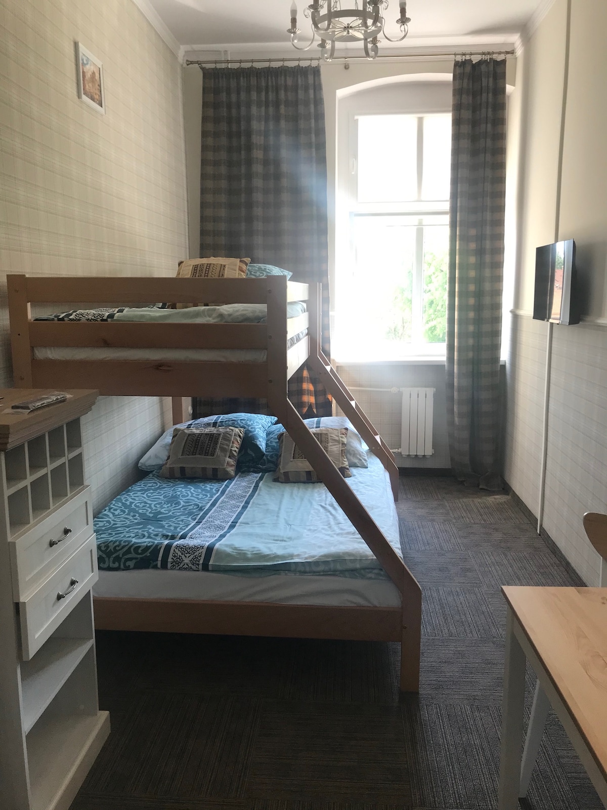 A cozy room features a wooden bunk bed with soft bedding and decorative pillows on the lower bed. A small desk and chair are present, along with a cabinet displaying storage options. Light enters through a window adorned with patterned curtains.
