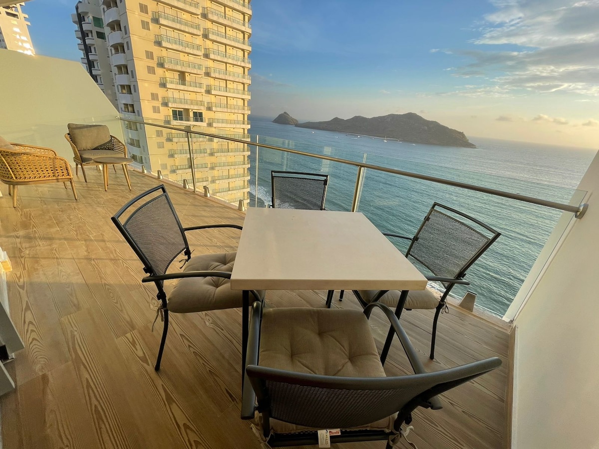 An outdoor terrace is shown, equipped with a dining table and four chairs, overlooking the ocean. The view includes distant islands and a serene sky at sunset, creating a peaceful ambiance for guests to enjoy.