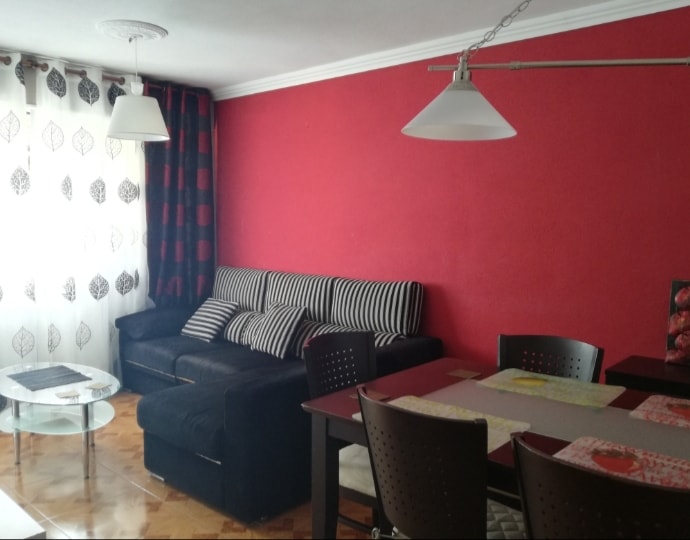 A cozy living and dining area is presented, featuring a black sofa adorned with striped cushions. A glass coffee table is positioned in front of the sofa, while a dark dining table with metallic chairs is set nearby. Bright red walls are complemented by patterned curtains.