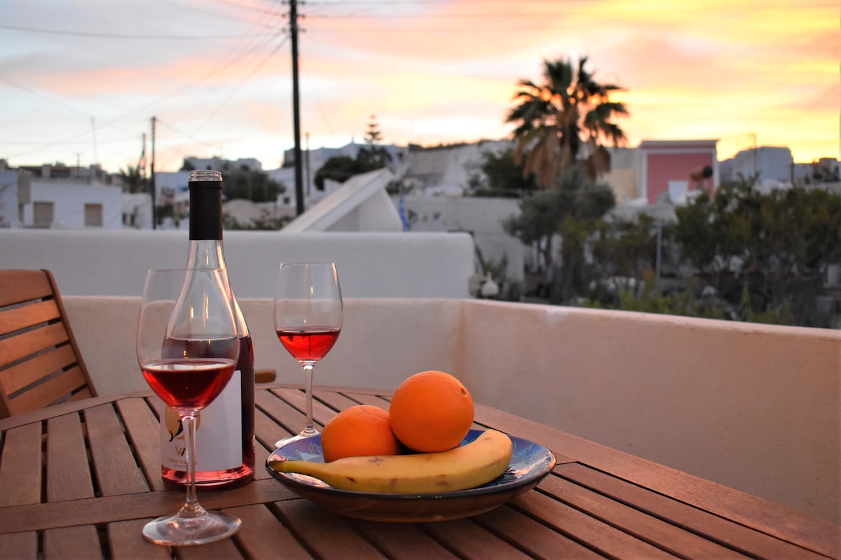 A wooden table is set with two glasses filled with rosé wine alongside a plate of fresh fruit, including oranges and a banana. A tranquil sunset paints the sky in soft hues behind the village's white buildings and palm trees.