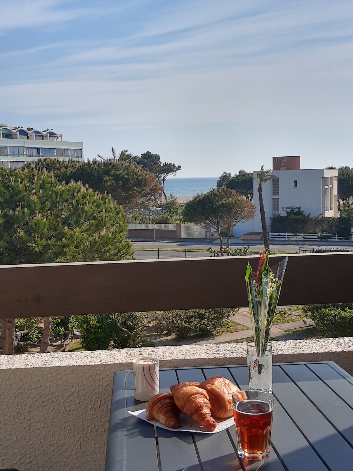 A terrace table is set with beverages and fresh croissants, overlooking a view of the sea. In the background, trees and nearby buildings are visible, contributing to a serene atmosphere. The sky is bright, enhancing the welcoming scene.