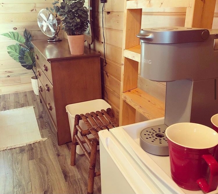 A cozy interior space features wooden walls and a light-colored floor. A small fan is positioned next to a potted plant atop a dresser, enhancing the natural feel. A compact coffee maker sits on a mini-fridge, accompanied by two red cups, ready for use.