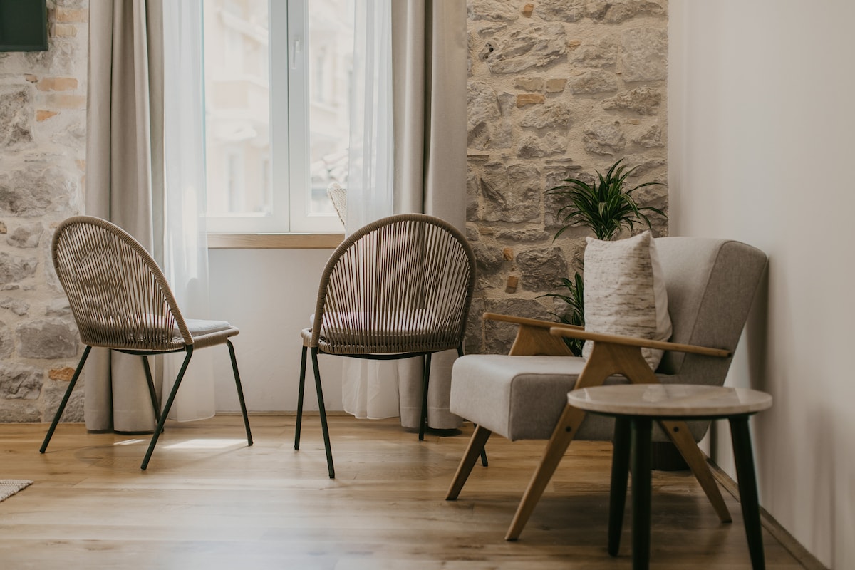 A cozy seating area features two rattan chairs and a modern armchair arranged around a small round table. Natural light enters through wide windows, accentuating the exposed stone wall and timber flooring.