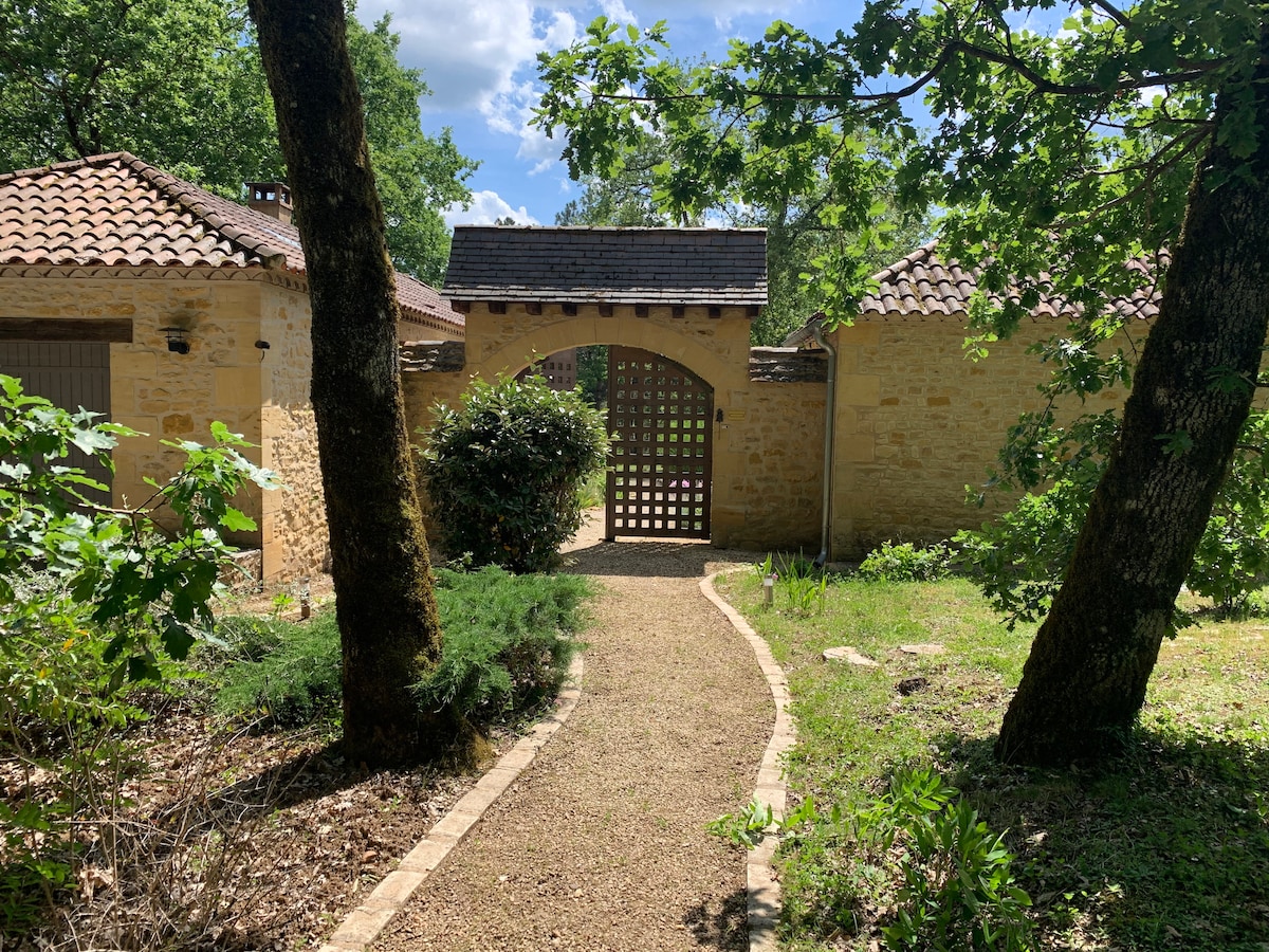 A winding gravel pathway leads to a charming wooden gate, framed by lush greenery and trees. The weathered stone walls of the surrounding buildings reflect a rustic character, while the bright sky enhances the inviting atmosphere of the property.