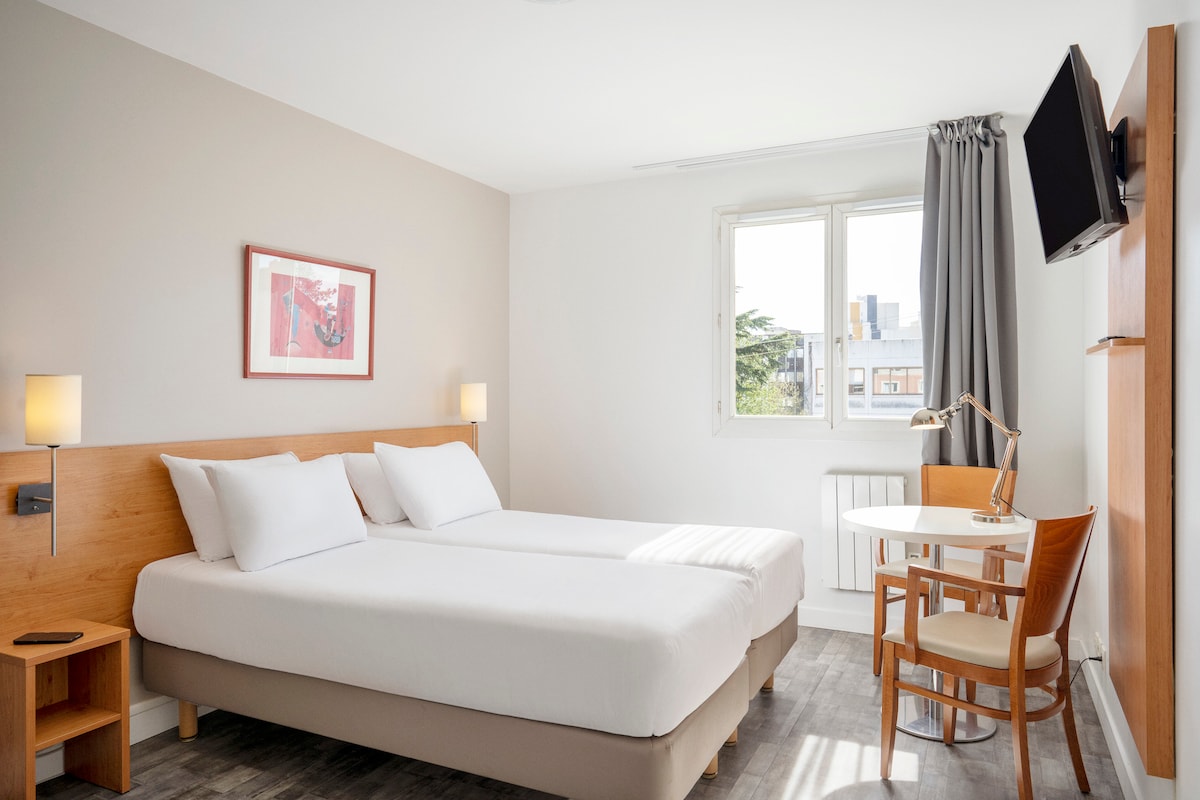 A modern room features a double bed with crisp white linens. Natural light enters through a large window, illuminating a small round table and a wooden chair. A wall-mounted television is positioned across from the bed, enhancing the functional layout of the space.