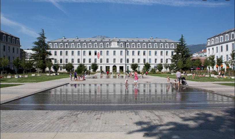 A spacious public square is depicted, featuring a large, shallow water basin reflecting the surrounding architecture. Families and children can be seen enjoying the water, with greenery lining the area and a backdrop of a historic building characterized by classic architecture and a clear blue sky.