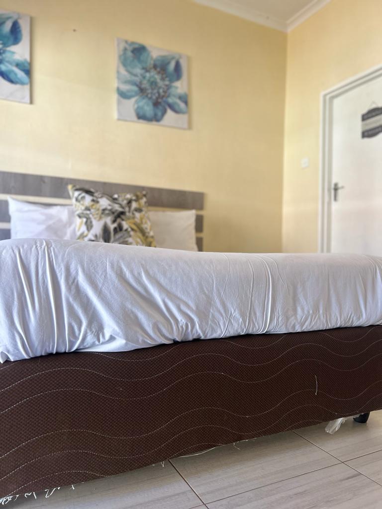 A spacious bed is the focal point of the room, with a neatly arranged white duvet cover. Soft decorative pillows add subtle comfort, while light yellow walls are complemented by floral artwork above the headboard, enhancing the room's inviting ambiance.