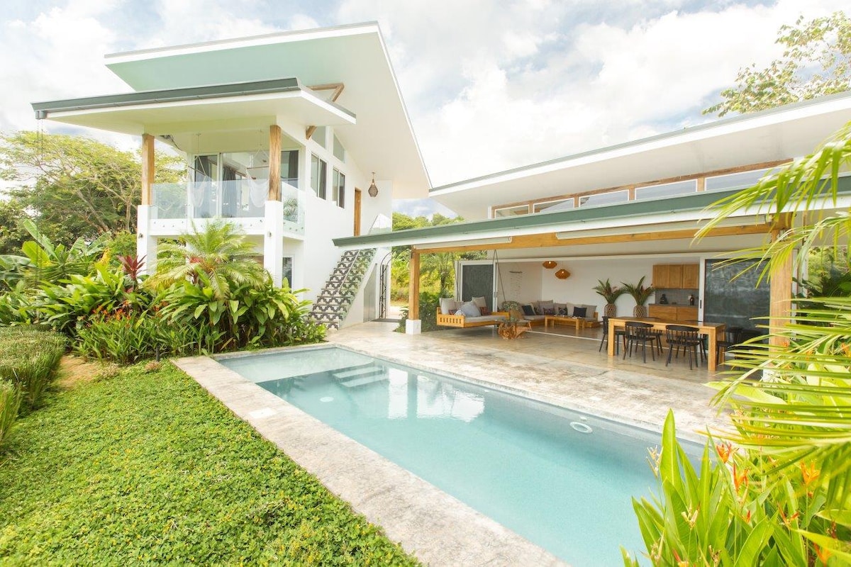 A modern vacation home is surrounded by lush greenery, featuring a private saltwater pool. The exterior showcases large glass sliding doors that open to an outdoor living area with seating. Sunlight filters through overhanging eaves, creating a bright and airy atmosphere.