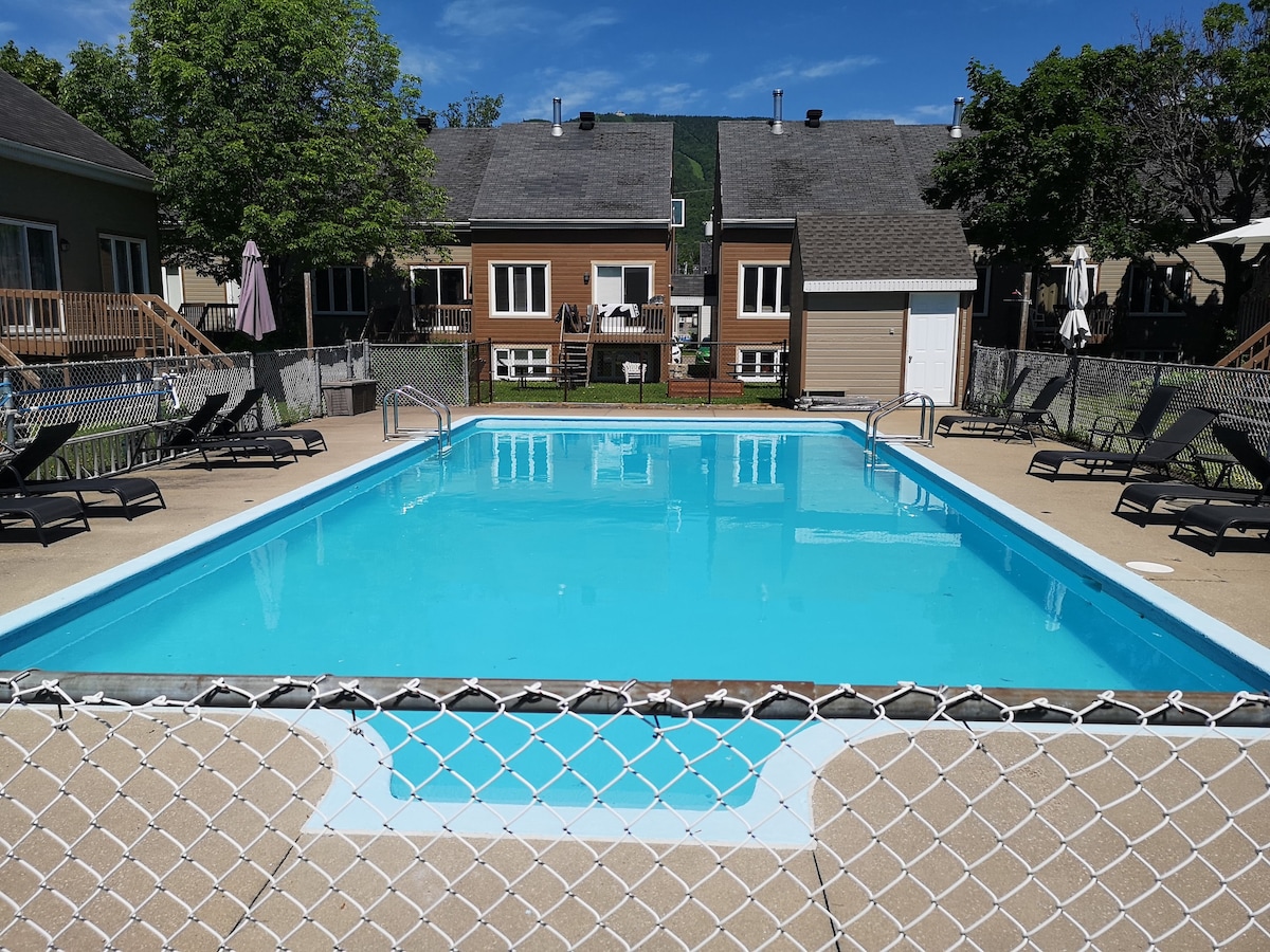 A clear, blue saltwater pool is surrounded by a safety fence, with multiple lounge chairs positioned along the edge. The area is framed by buildings featuring earthy tones, and lush greenery is visible in the background under a bright blue sky.